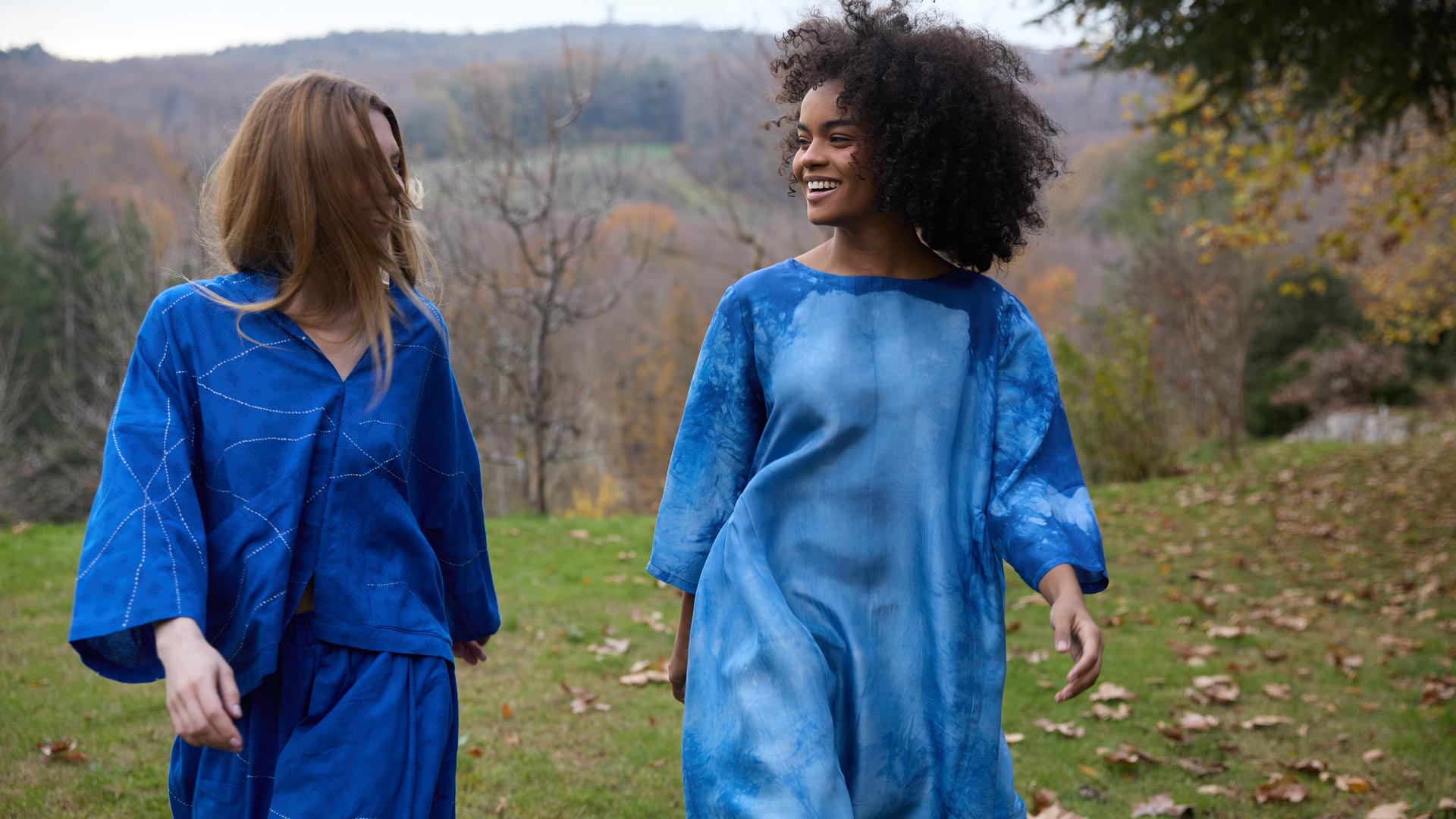 Two women walking through a grassy outdoor field wearing monochromatic blue garments. One wears a handwoven blue top with fine geometric Kantha stitching and matching trousers, while the other smiles wearing a flowing blue tie-dye maxi silk dress with cloud-like patterns.