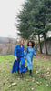 Two women walking through a grassy outdoor field wearing monochromatic blue garments. One wears a handwoven blue top with fine geometric Kantha stitching and matching trousers, while the other smiles wearing a flowing blue tie-dye maxi dress with cloud-like patterns.